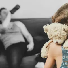 Dziewczynka z misiem na ramieniu, w tle niewyraźny mężczyzna pijący alkohol.