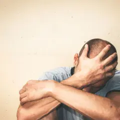 Mężczyzna zakrywa twarz dłońmi, siedząc przy jasnej ścianie.