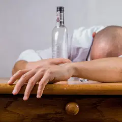 Mężczyzna śpi na stole, obok pusta butelka. Jego ręka opiera się o blat, symbolizując skutki nadmiernego spożycia alkoholu.