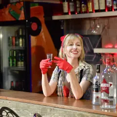 Uśmiechnięta barmanka w czerwonych rękawiczkach trzyma kieliszek z wódką. Obok stoją butelki, w tym duża 250 ml wódki.