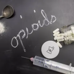Napis "opioidy" na tablicy, obok łyżeczki z proszkiem, strzykawki i butelki z tabletkami.