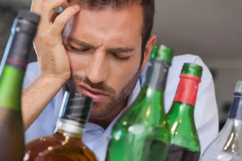 Mężczyzna z ręką na czole, otoczony butelkami. Wygląda na to, że wie, co to kac.