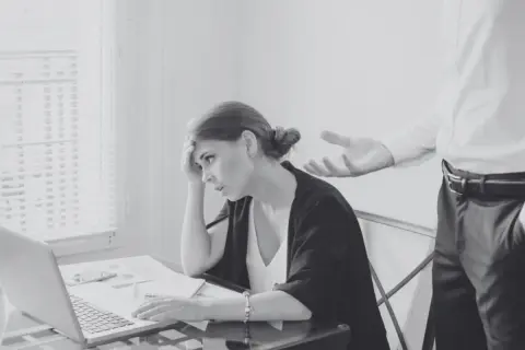 Kobieta z ręką na czole siedzi przy biurku z laptopem, obok niej stoi mężczyzna.