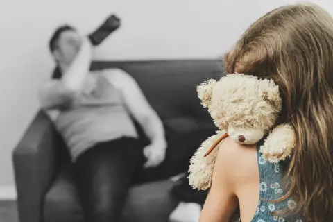 Dziewczynka z misiem na ramieniu, w tle niewyraźny mężczyzna pijący alkohol.