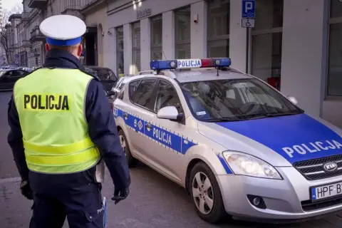 Policjant w kamizelce odblaskowej stoi obok radiowozu z napisem "POLICJA".