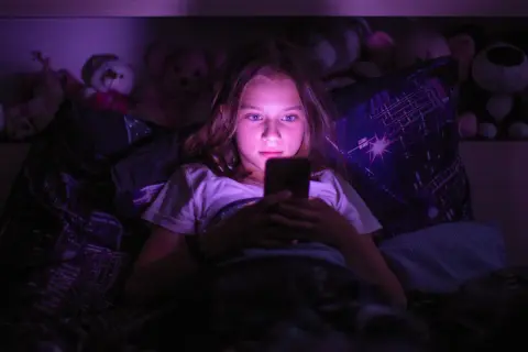 Dziewczynka leżąca w łóżku, patrząca w ekran telefonu w ciemności.