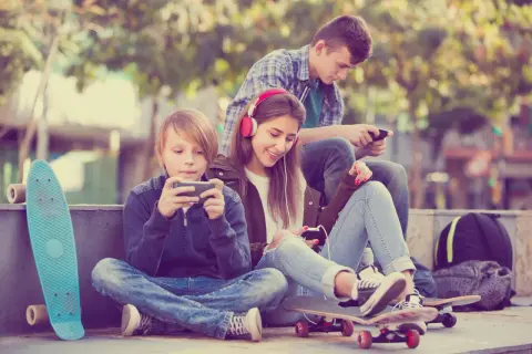 Trójka nastolatków pochłonięta smartfonami, obok deskorolki. Czy to początek uzależnień behawioralnych?