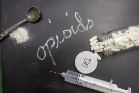 Napis "opioidy" na tablicy, obok łyżeczki z proszkiem, strzykawki i butelki z tabletkami.