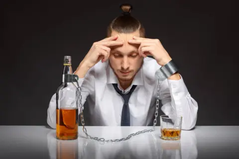 Mężczyzna w kajdanach, przykuty do butelki i szklanki z alkoholem, symbolizuje walkę z nałogiem. Suchy alkoholik, który mimo abstynencji, wciąż czuje ciężar uzależnienia.