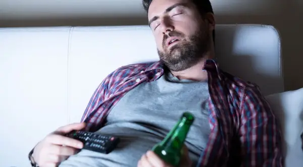 Mężczyzna śpi na kanapie z pilotem i butelką piwa, być może doświadczając dziwnych snów po odstawieniu alkoholu.
