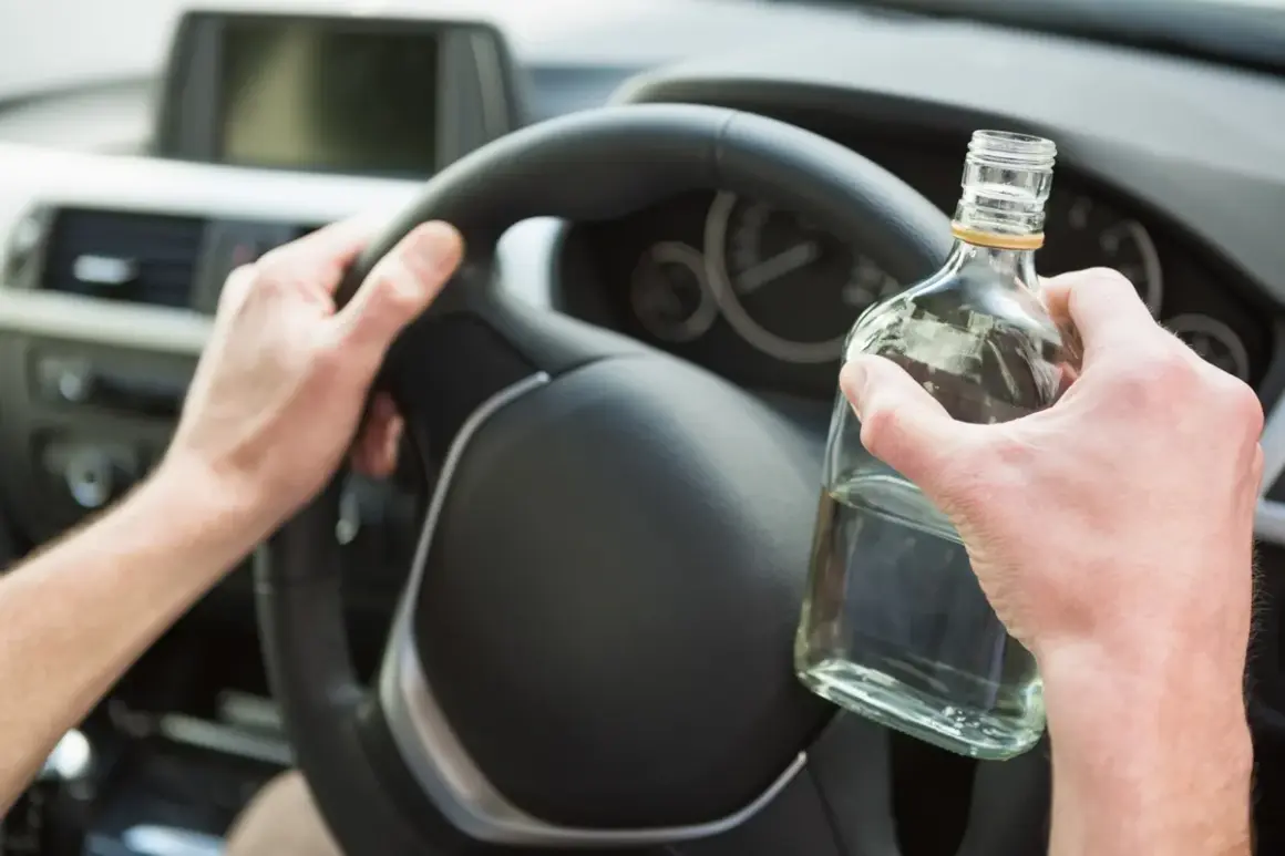Ręce kierowcy na kierownicy, jedna trzyma butelkę alkoholu. To niebezpieczne zachowanie, które łamie limit alkoholu w Polsce.