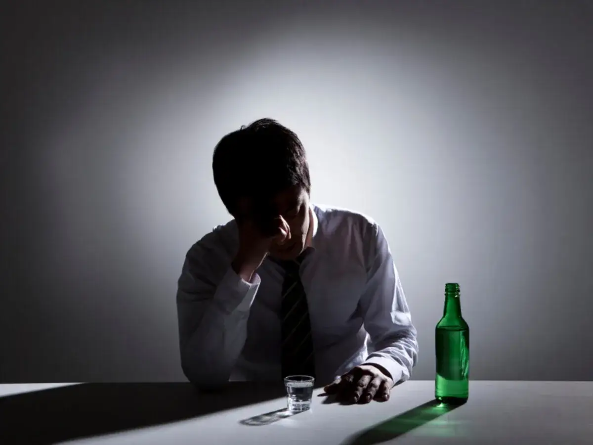 Mężczyzna w białej koszuli i krawacie, ze smutną miną, siedzi przy stole z butelką zielonego alkoholu i kieliszkiem. Jak rozpoznać chemola?