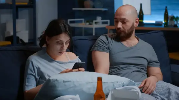 Para siedzi na kanapie, kobieta patrzy w telefon, mężczyzna obok niej wygląda na zamyślonego.