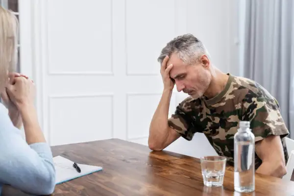Mężczyzna w mundurze maskującym, z ręką na czole, przeżywa trudne chwile. Rozmowa terapeutyczna, być może przymusowe leczenie alkoholika, jest jego jedyną nadzieją.