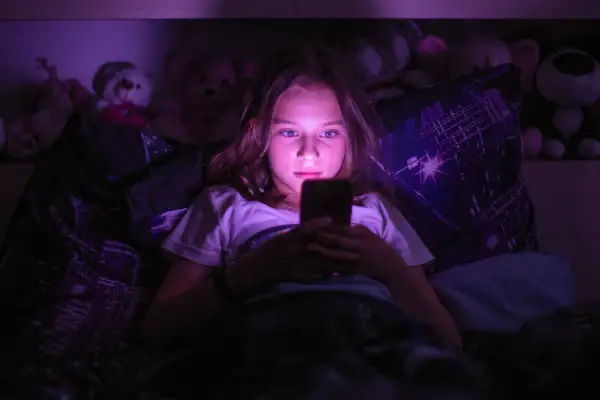 Dziewczynka leżąca w łóżku, patrząca w ekran telefonu w ciemności.