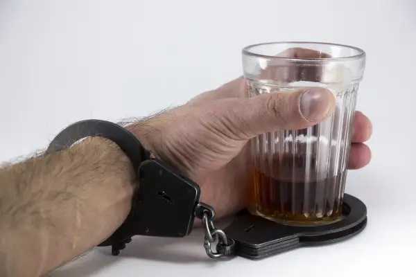 Ręka w kajdankach trzyma szklankę z alkoholem. Symbolizuje to problem, co zrobić z alkoholikiem awanturnikiem, który jest uwięziony w nałogu.
