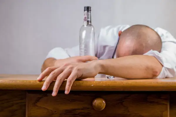 Mężczyzna w białej koszuli leży na stole, głowa oparta na ramieniu. Obok pusta butelka, znak przedawkowania alkoholu.
