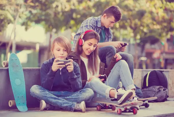 Trójka nastolatków pochłonięta smartfonami, obok deskorolki. Czy to początek uzależnień behawioralnych?
