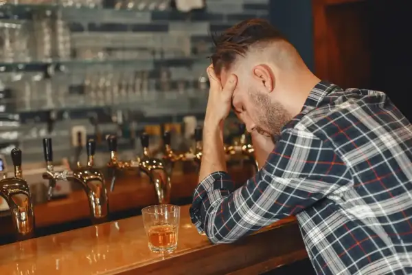 Mężczyzna z rękami na głowie siedzi przy barze, obok szklanka z alkoholem. Może to być jeden z 6 objawów uzależnienia od alkoholu.